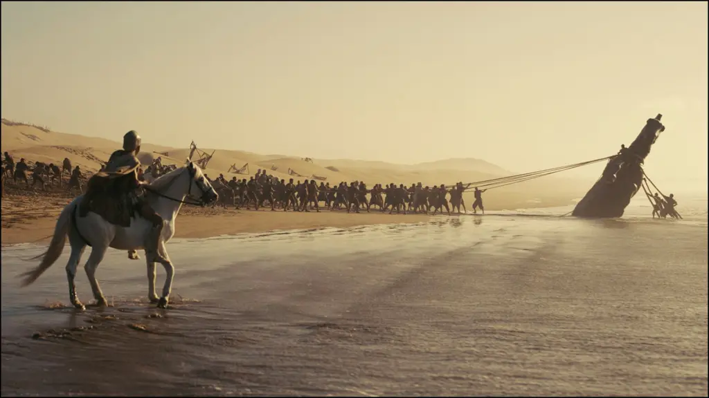 Hundreds of people and a rider on horseback pull a massive shipwreck ashore on a sandy beach at sunset, using long ropes.